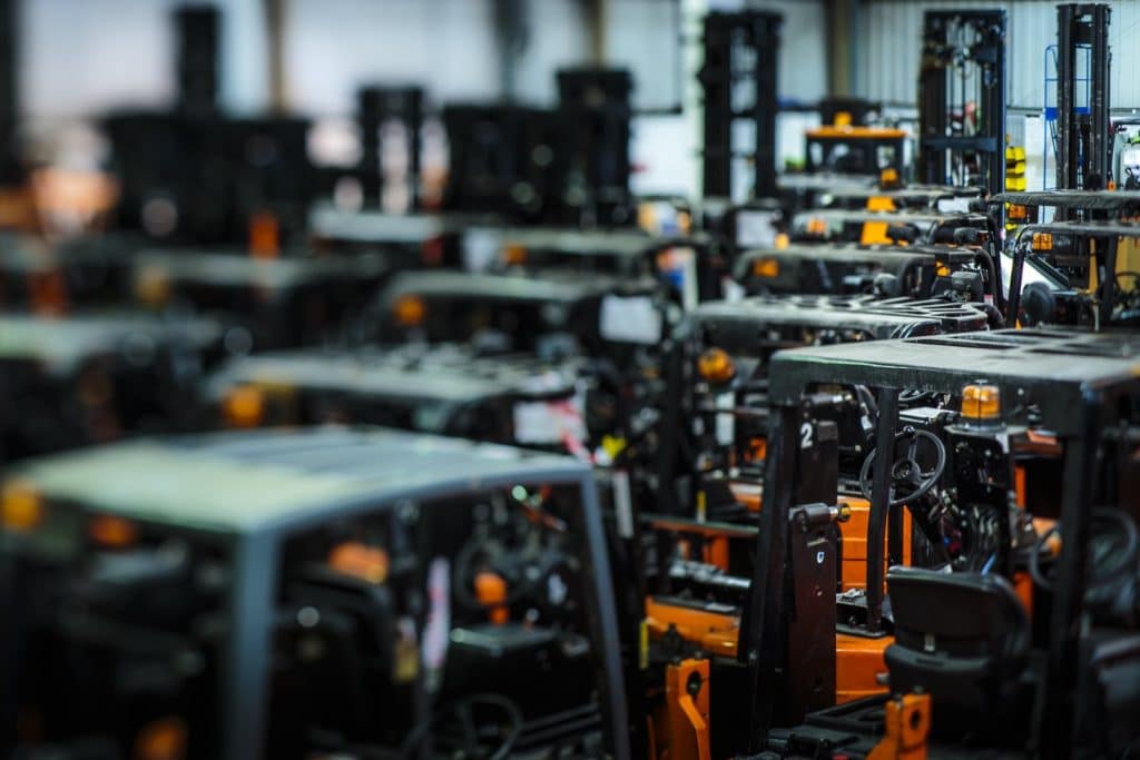 A bustling warehouse showcases a collection of forklifts available for hire, closely arranged together. The image captures the dense placement, with a shallow depth of field highlighting one in the foreground while others are subtly blurred in the background, emphasizing their organized alignment.