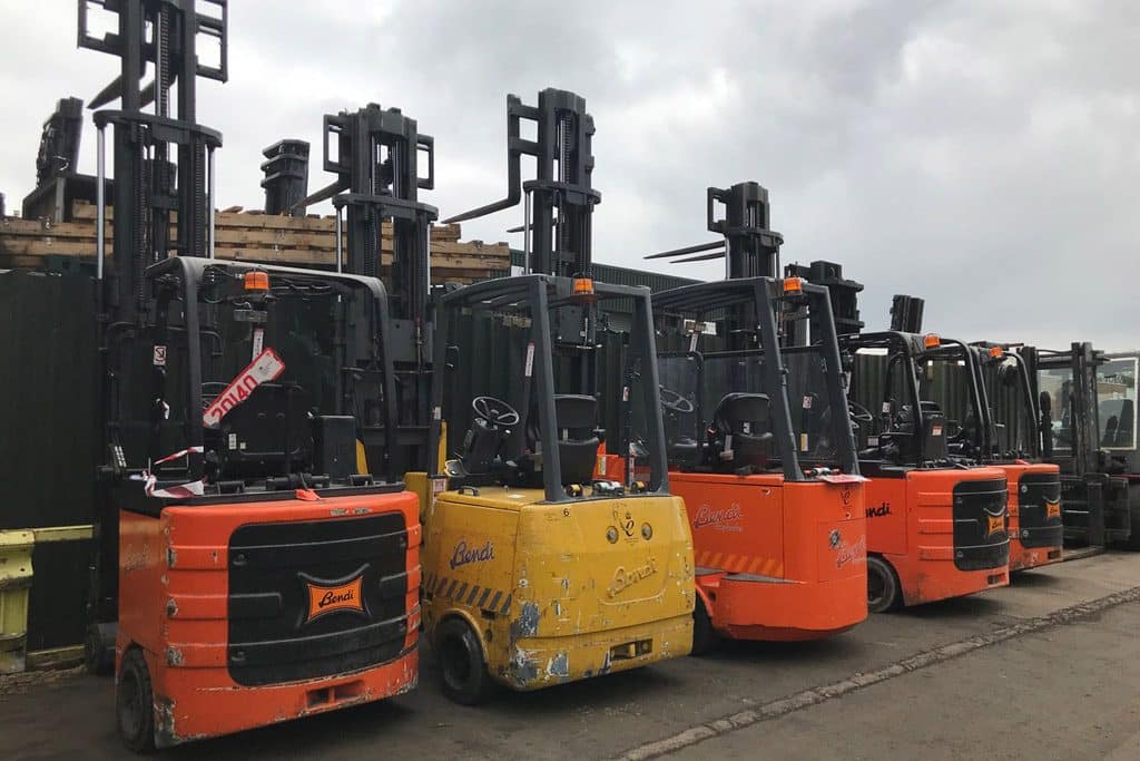 A row of five used forklifts, primarily orange in color, lined up outdoors against a green fence. The overcast sky creates a gray backdrop.