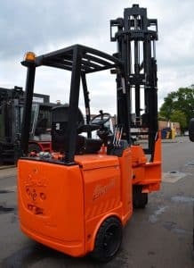 A refurbished Bendi B40, colored in orange and equipped with a black overhead guard, is stationed on a paved surface. Its forks are elevated against the setting of another forklift and industrial structures under a cloudy sky.