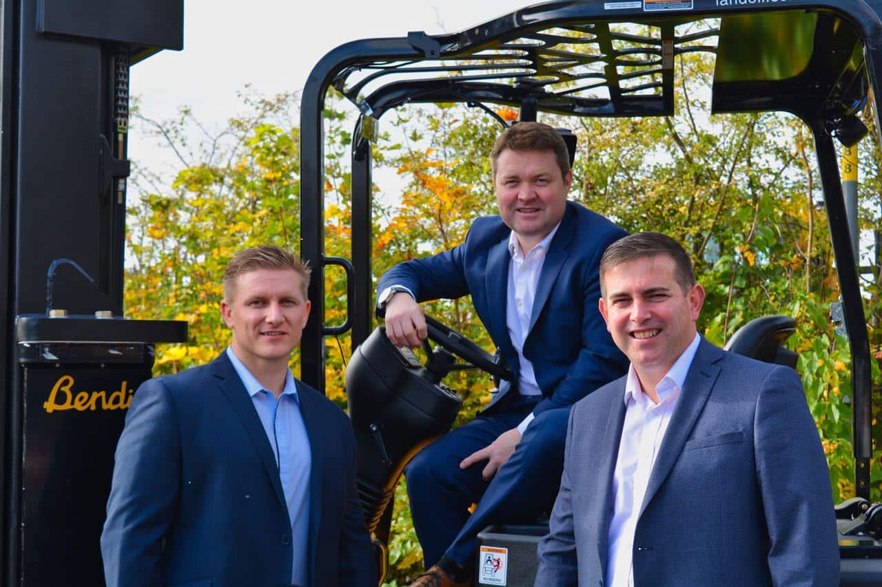 Three men in suits, part of senior management, are posing together outdoors. One is seated at the controls of a large Translift machine, while the other two stand nearby, all smiling. Trees with autumn foliage form a vibrant background.