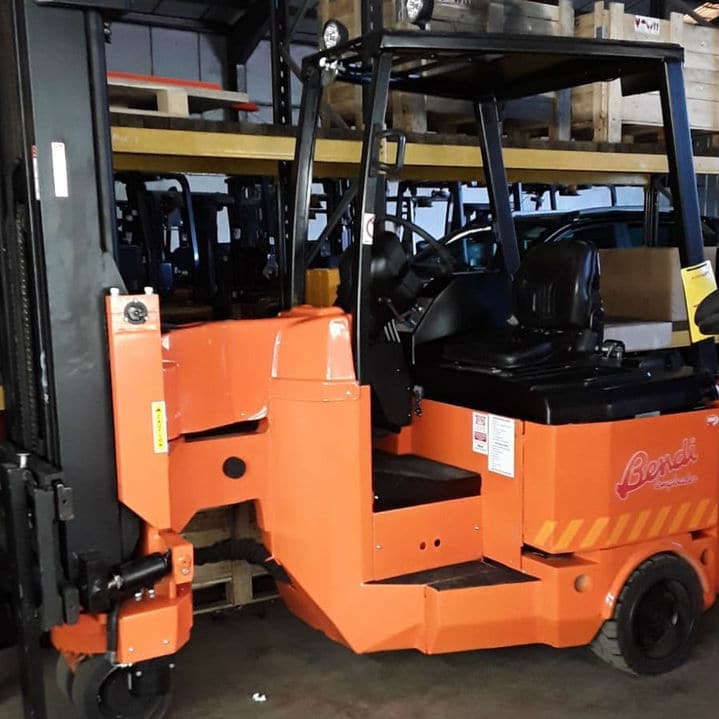 An orange, refurbished Bendi Longloader is parked indoors, proudly displaying its logo. In the background of this warehouse scene, shelving and multiple pallets are visible, creating a busy industrial atmosphere.