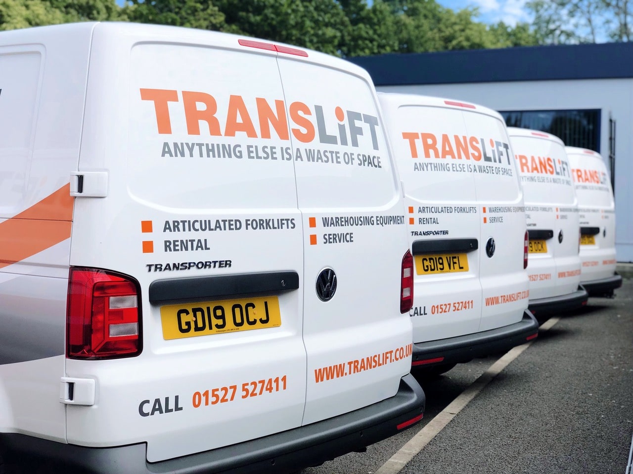 A row of white vans with large TRANSLIFT logos parked side by side. The vans, signaling exciting times ahead, advertise services like articulated forklifts, rental, and warehousing equipment. Contact details and a phone number are prominently displayed on the back of the vans.