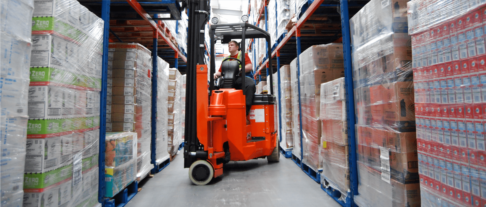 In the bustling warehouse, a worker skillfully maneuvers a Translift forklift down an aisle lined with well-organized shelves, efficiently transporting a load amid towering stacks of packaged goods.