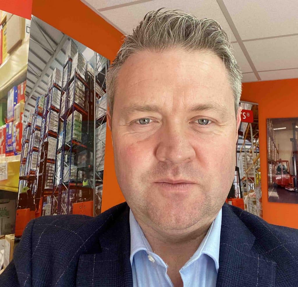 A man with short grey hair is taking a selfie indoors, likely representing the Clean Growth Team. He wears a dark suit jacket over a light blue shirt. In the background, high stacks of boxes and shelves indicate a warehouse setting with an orange wall.