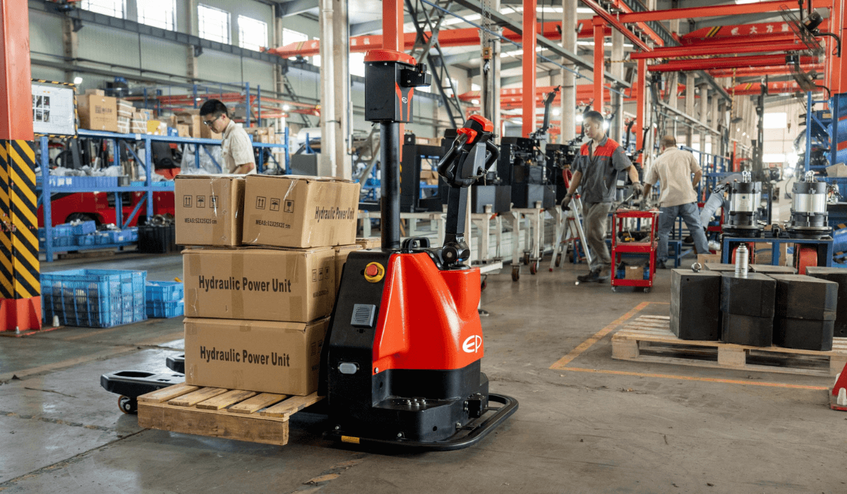 In a bustling factory setting, workers expertly operate machinery. Amidst the activity, a red electric pallet jack from EP Equipment efficiently transports boxes labeled Hydraulic Power Unit, with various equipment and busy workers in the background.
