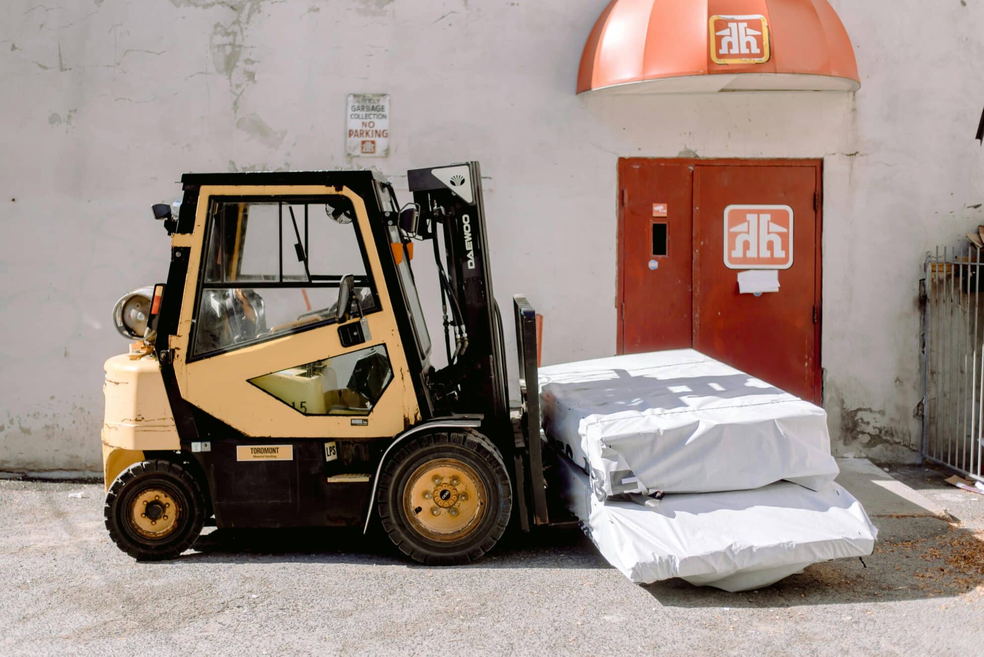 A forklift hire service efficiently transports wrapped packages in front of a building with a red door and an orange awning. A No Parking sign is visible on the wall, while the skilled forklift operator maneuvers the vehicle with precision.