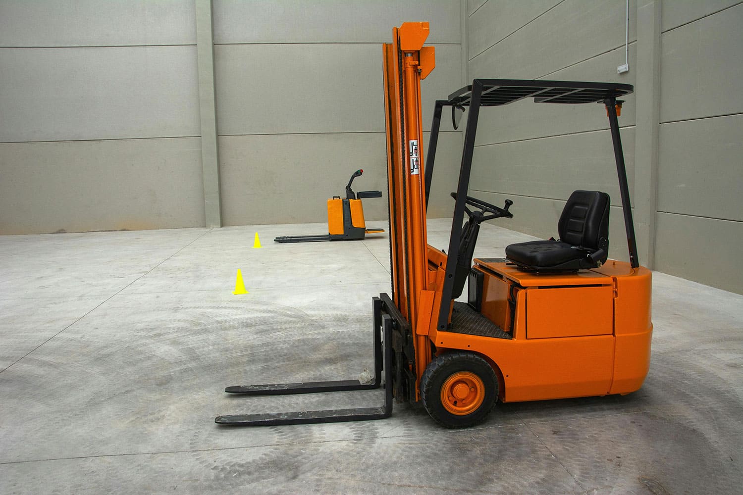 An orange forklift is parked in a spacious, empty warehouse with two yellow cones near another forklift, showcasing an affordable solution for efficient operations. The gray floor and walls highlight the clean, organized environment ideal for smooth workflow.