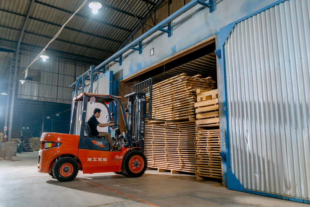A person operating a red forklift is expertly moving a large stack of wooden planks into a warehouse with tall, open sliding doors. The interior, industrial with high ceilings and overhead lights, showcases why EP Forklifts are the best choice for efficient business operations.