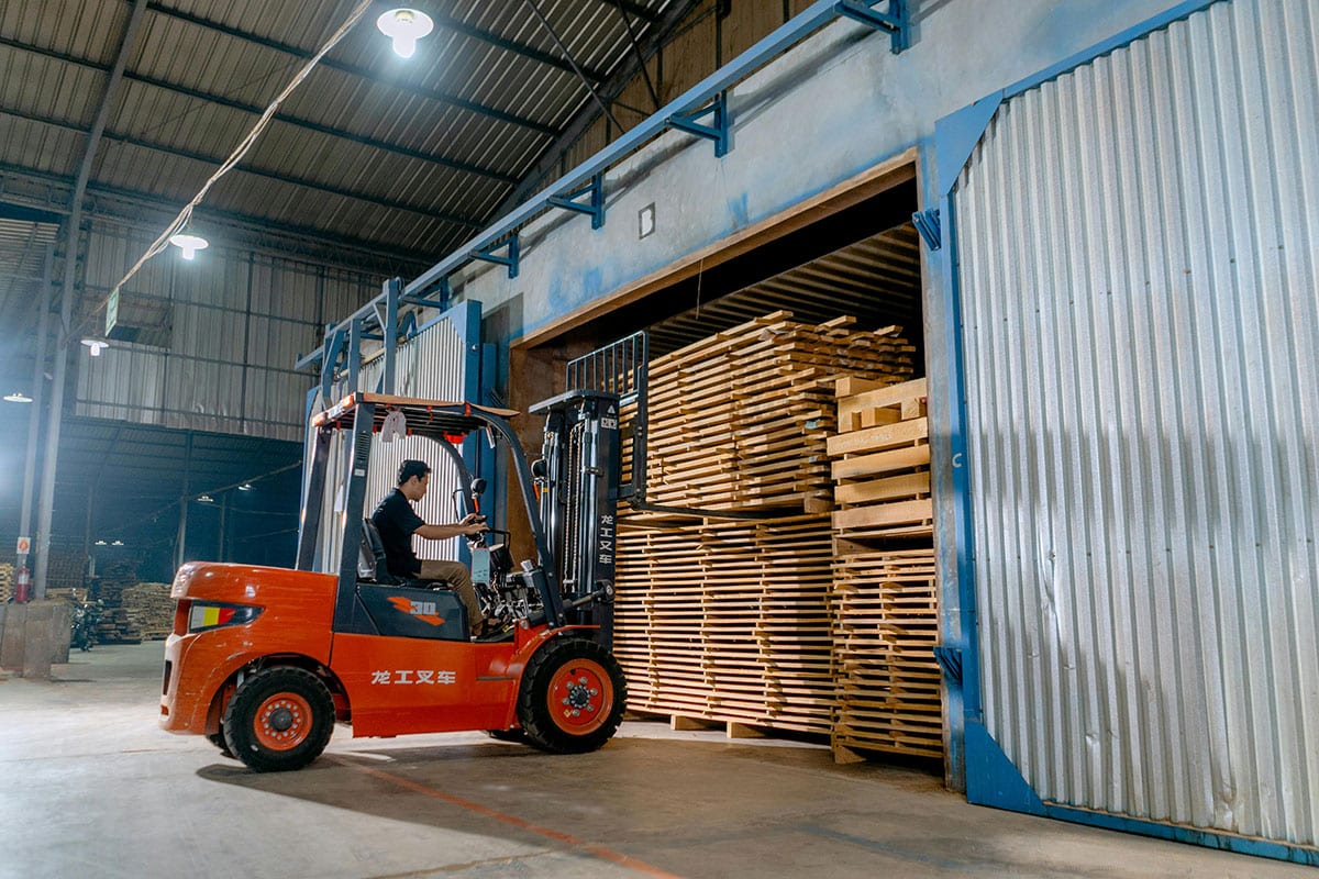 A person operating a red forklift is expertly moving a large stack of wooden planks into a warehouse with tall, open sliding doors. The interior, industrial with high ceilings and overhead lights, showcases why EP Forklifts are the best choice for efficient business operations.