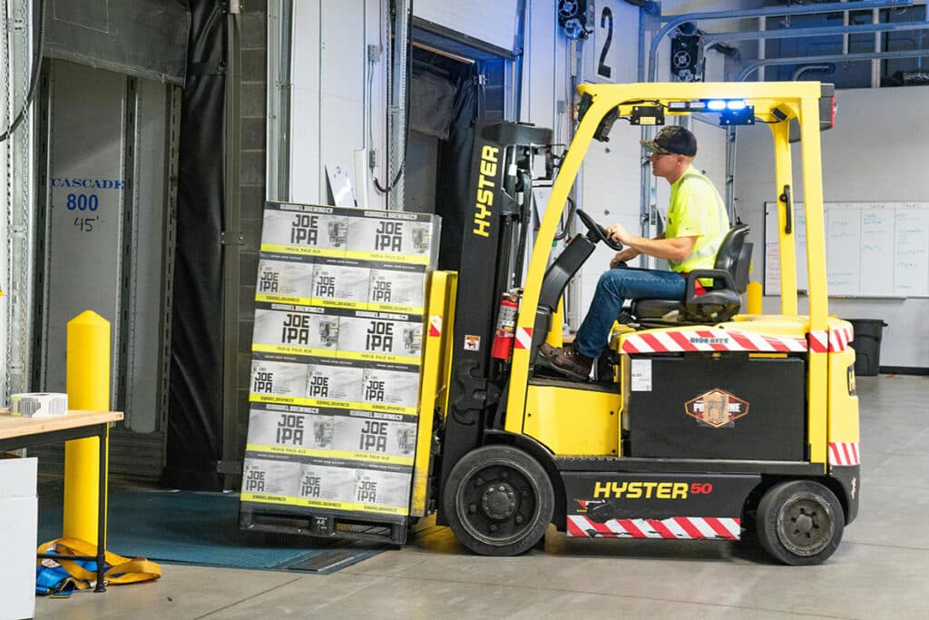 A person skillfully maneuvers a yellow articulated forklift truck, moving several boxes labeled Joe IPA into the building. The setting appears to be a warehouse with open dock doors and visible safety markings, efficiently utilizing space savers to maximize storage capacity.