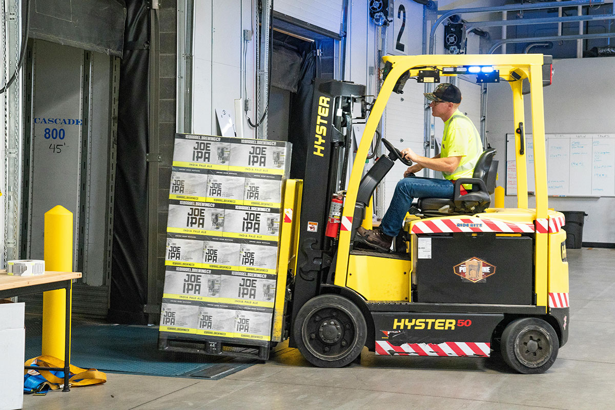 A person skillfully maneuvers a yellow articulated forklift truck, moving several boxes labeled Joe IPA into the building. The setting appears to be a warehouse with open dock doors and visible safety markings, efficiently utilizing space savers to maximize storage capacity.