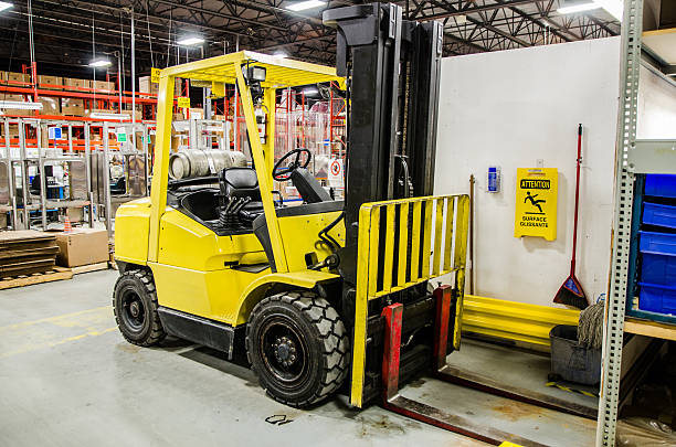A yellow VNA forklift is parked in an industrial warehouse. The machine is near a wall with a caution sign and shelving filled with various items. The environment appears organized, with pallets and equipment visible in the background.