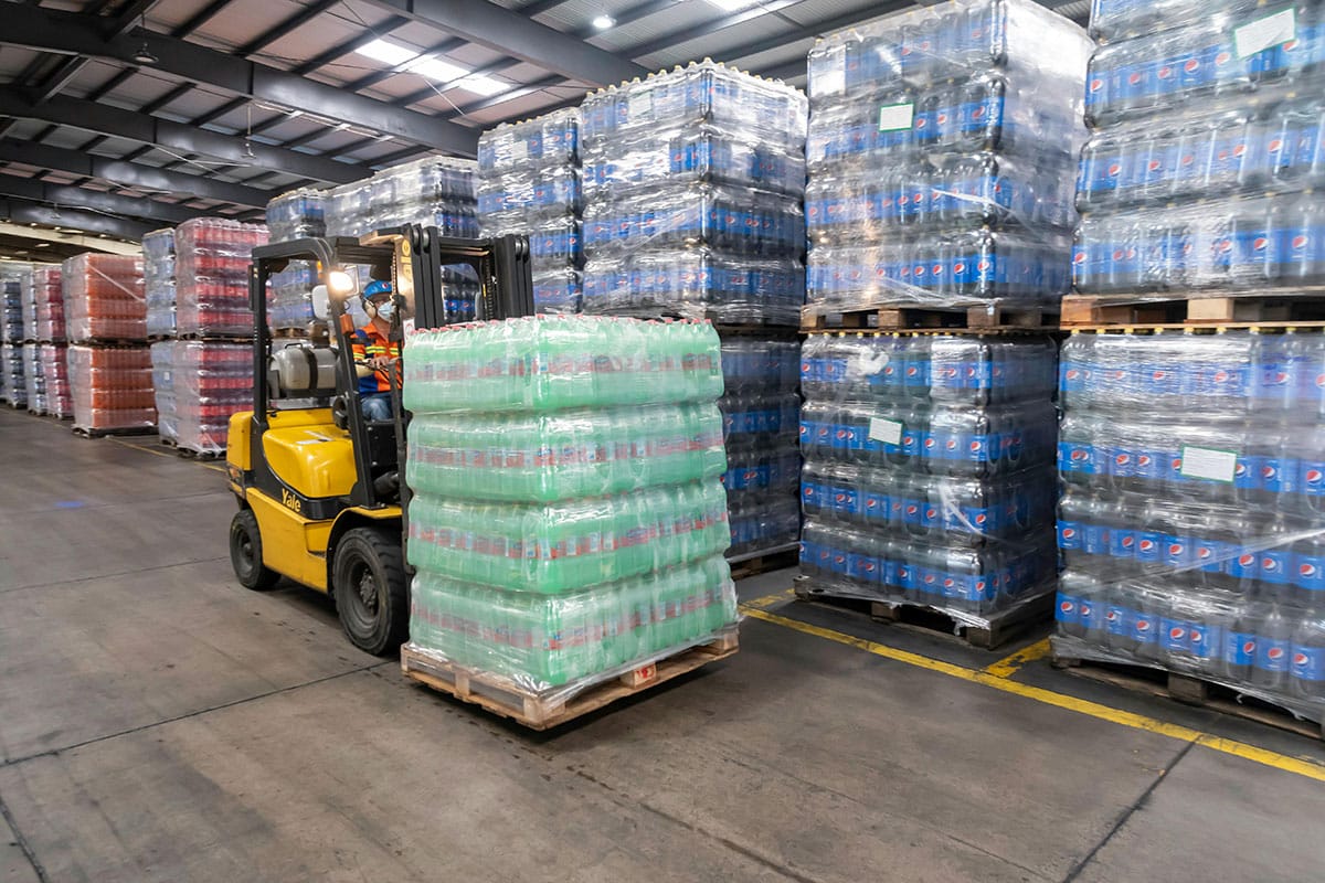 An articulated forklift truck transports a pallet of bottled beverages wrapped in clear plastic in a warehouse. Stacks of similar pallets are visible in the background. The space, illuminated by overhead lighting, features concrete floors.