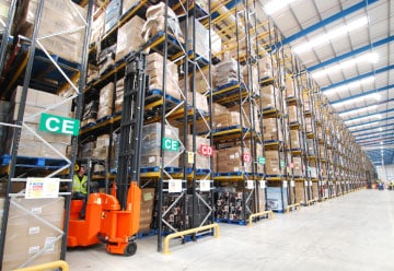 A warehouse interior with tall racks filled with stacked boxes reminiscent of a B&M Bargains setup. An orange forklift is lifting a pallet on the left side, operated by a person in a high-visibility vest. Overhead lighting and signs mark different sections like CE and CD.