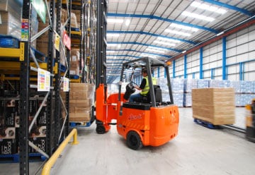 An orange forklift, operated by a person in a high-visibility vest, is moving a pallet of boxes in the well-lit B&M warehouse with tall shelves. Other boxed items are visible on the shelves and in the background, showcasing the vast array of bargains available at B&M.