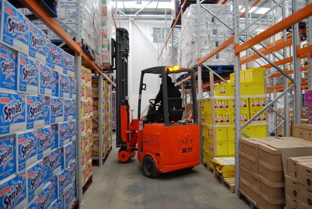 A forklift operates in a bustling warehouse aisle stacked with colorful packaged goods for Bobby's Food on either side. The shelves are filled with boxes destined for Bobby’s Restaurant, and a worker is visible driving the bright orange forklift.
