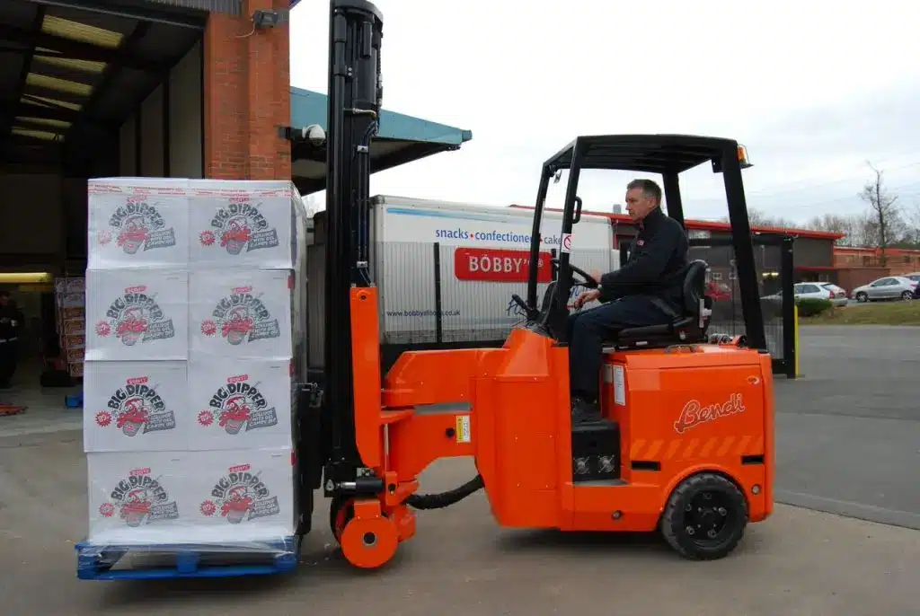 A man skillfully operates an orange Bendi forklift, moving a pallet loaded with Bobby's Food-branded boxes. The forklift exits a warehouse, with a truck visible in the background of this bustling industrial area.