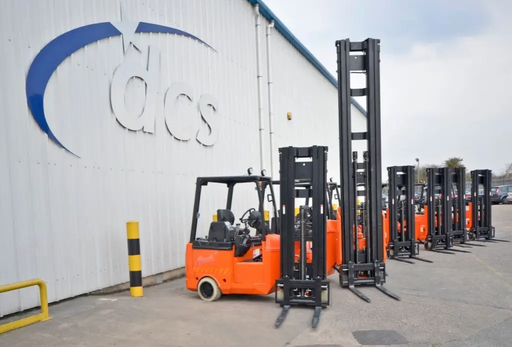 Five orange forklifts are lined up outside a building with a large DCS Group sign on the wall. The area is paved, and the UK sky is overcast, adding an industrial charm to the scene.