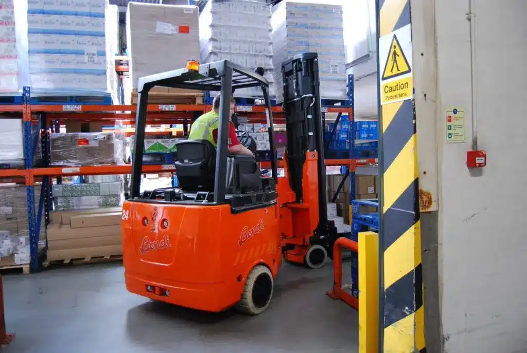 In a UK warehouse, a worker skillfully operates an orange forklift, maneuvering between shelves filled with boxes. A yellow caution sign stands nearby as the DCS Group stocks the space with various packaged goods and equipment.