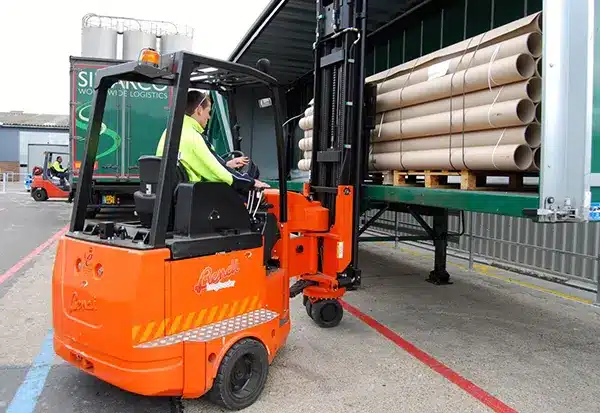A person operates an orange forklift with dual functionality, loading long loads of cylindrical pipes onto a truck at a logistics facility. The truck is partially covered, and the worker is dressed in a high-visibility jacket. The scene unfolds in an industrial area.