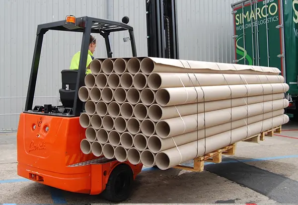 A worker operates an orange forklift designed for dual functionality, effortlessly handling long loads of stacked cardboard tubes secured on a pallet. The scene unfolds in an industrial area, where partially visible green Fiberweb logistics signage decorates the background.