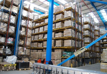 A large warehouse filled with stacked pallets on tall shelves, organized in multiple rows. The area is spacious with visible steel beams and lighting from the ceiling. Amidst it all, boxes labeled "Harris Brushes" stand ready on a conveyor belt in the foreground.
