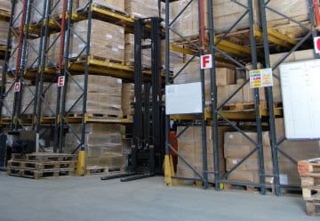 A forklift accesses a pallet in a warehouse aisle, navigating between tall industrial shelves filled with stacked boxes of Harris Brushes and paintbrushes. Signs with letters and notices are visible on the shelves. The floor is clear, keeping the painting tools organized and accessible.