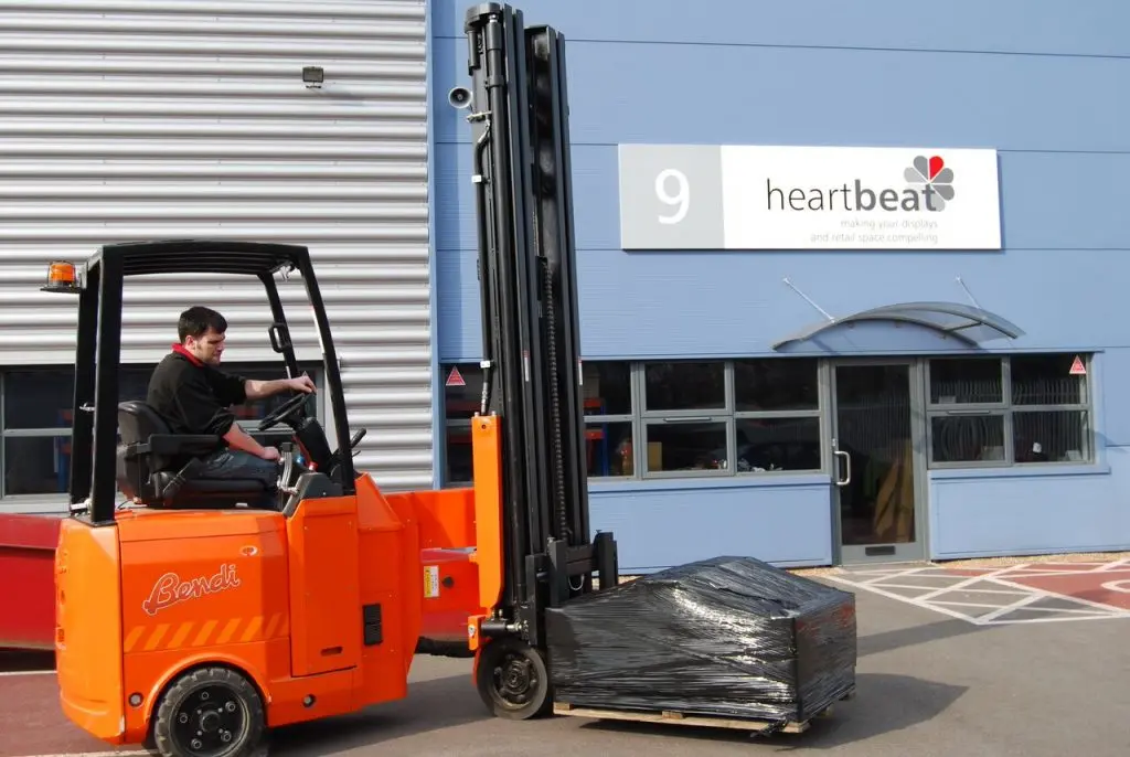 A person operating an orange forklift is lifting a wrapped pallet outside a building marked with the number 9. The sign on the building reads Heartbeat Manufacturing, suggesting it’s part of an extensive industrial complex. The area is neatly paved, enhancing the facility's organized appearance.