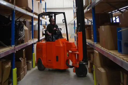 A man operates an orange forklift in a bustling warehouse aisle, surrounded by shelves stacked with various boxes and packages. The high-ceilinged facility, a hub of Heartbeat Manufacturing, is illuminated with both natural and artificial light.