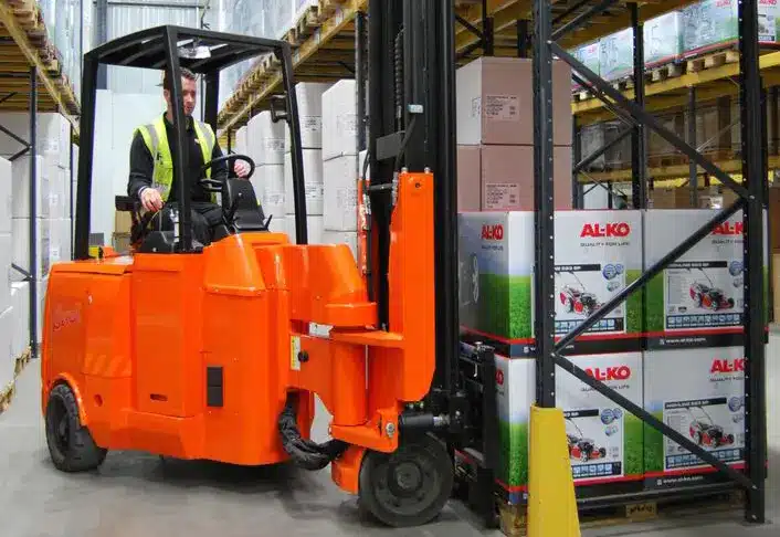 A person operates an orange forklift, deftly maneuvering it to lift and organize boxes labeled AL-KO. The scene unfolds in a warehouse filled with tall shelves holding various items related to Garden Machinery. Clad in a high-visibility vest, the worker navigates the busy Rochford facility.