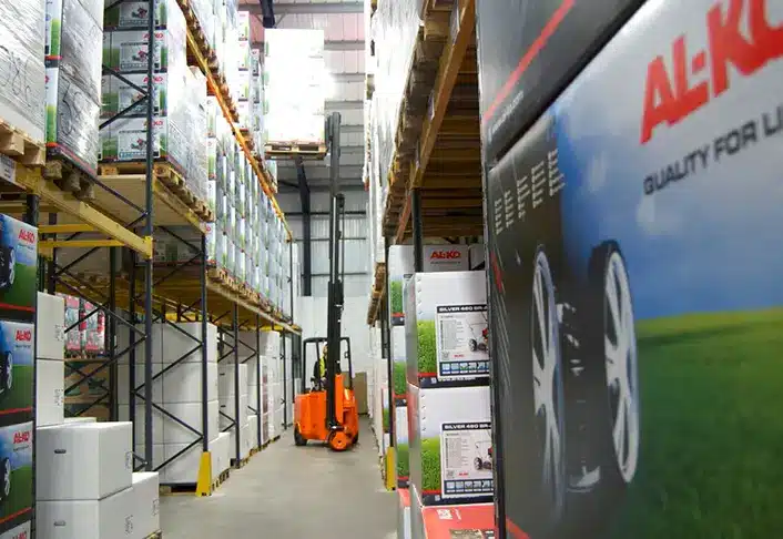 A well-organized warehouse interior with stacked shelves filled with boxes. A bright orange forklift, a vital piece of machinery, is parked in the aisle. Boxes display the brand AL-KO and feature images of wheels. The space is efficiently arranged and brightly lit, reminiscent of Rochford's precision.