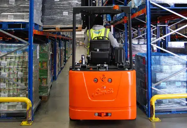 A warehouse worker in a reflective vest operates an orange reach truck, skillfully navigating narrow aisles lined with stacked pallets of Symington’s Foods on blue metal shelves.