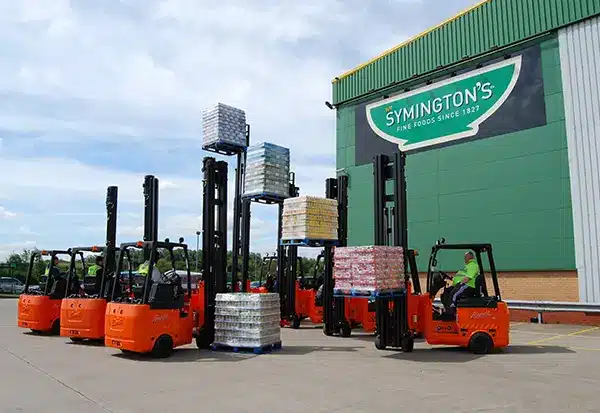 A group of orange forklifts operates in an outdoor warehouse, lifting pallets stacked with various goods. The building displays a large sign reading "Symington’s Foods Since 1827." Workers are seated on the forklifts under a partly cloudy sky, efficiently managing the inventory for Symington’s Foods.