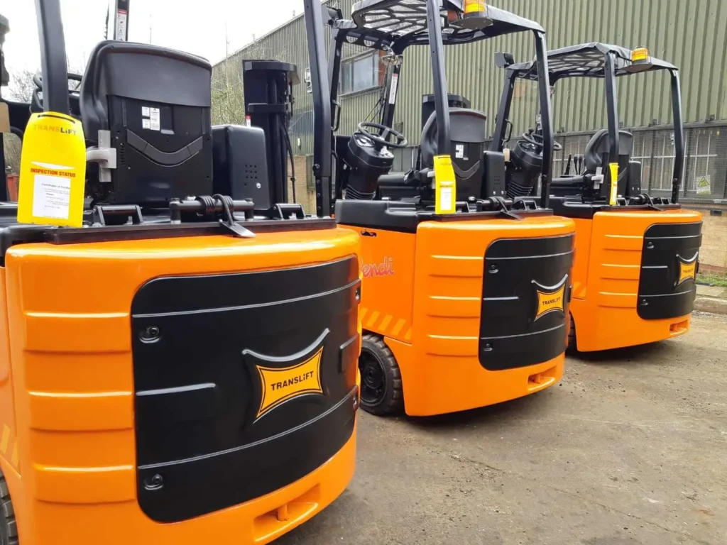 Three orange and black forklifts, maintained by Forklift Specialists, are lined up outdoors, showing the front view. They are parked beside a large industrial building with green corrugated walls. Each forklift has a yellow tag hanging on it.